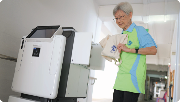 An older woman loads a cloth bag into an open compartment of a white delivery robot in a hallway.
